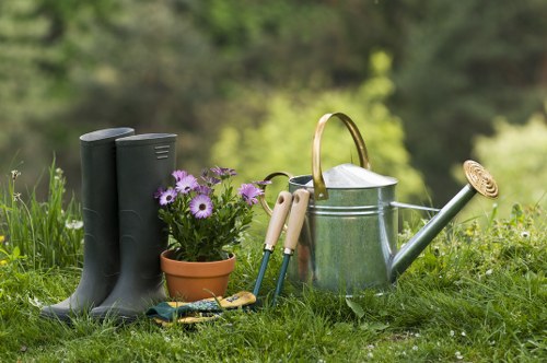 The image displays a neatly maintained garden scene with various gardening tools and elements arranged on a grassy lawn. In the foreground, a pair of black rubber gardening boots stand upright next to a small terracotta pot filled with purple and white daisy-like flowers, adding a touch of colour to the scene. Nearby, a set of garden hand tools, including pruning shears and a trowel with cream handles, are placed on the grass, indicating recent or ongoing garden work. A metallic watering can with a curved spout and a wooden handle is positioned beside the tools, ready for watering plants. The background reveals lush greenery with blurred trees and foliage, suggesting a peaceful outdoor space typical of a residential garden in Waterloo. The garden surface exhibits a mixture of vibrant green grass and patches of exposed soil or paving, with natural light illuminating the scene under an overcast or partly cloudy sky, evoking a calm and well-maintained outdoor environment suitable for gardening services from Hedge Trimming Waterloo.