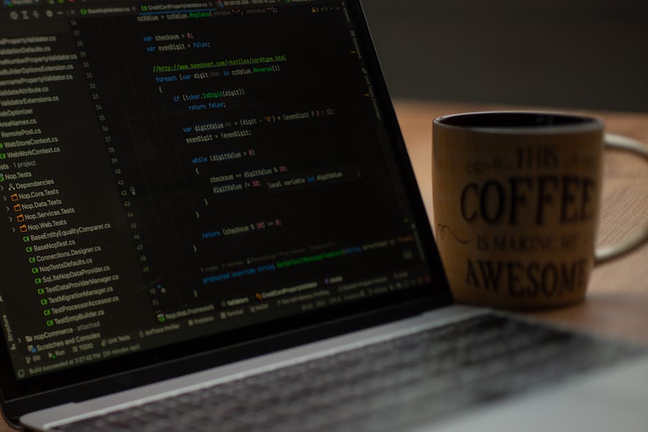 A person's hands typing on a sleek laptop keyboard, displaying lines of green and blue code on the screen in a dark-themed programming interface. The setting appears to be indoors, with the laptop res